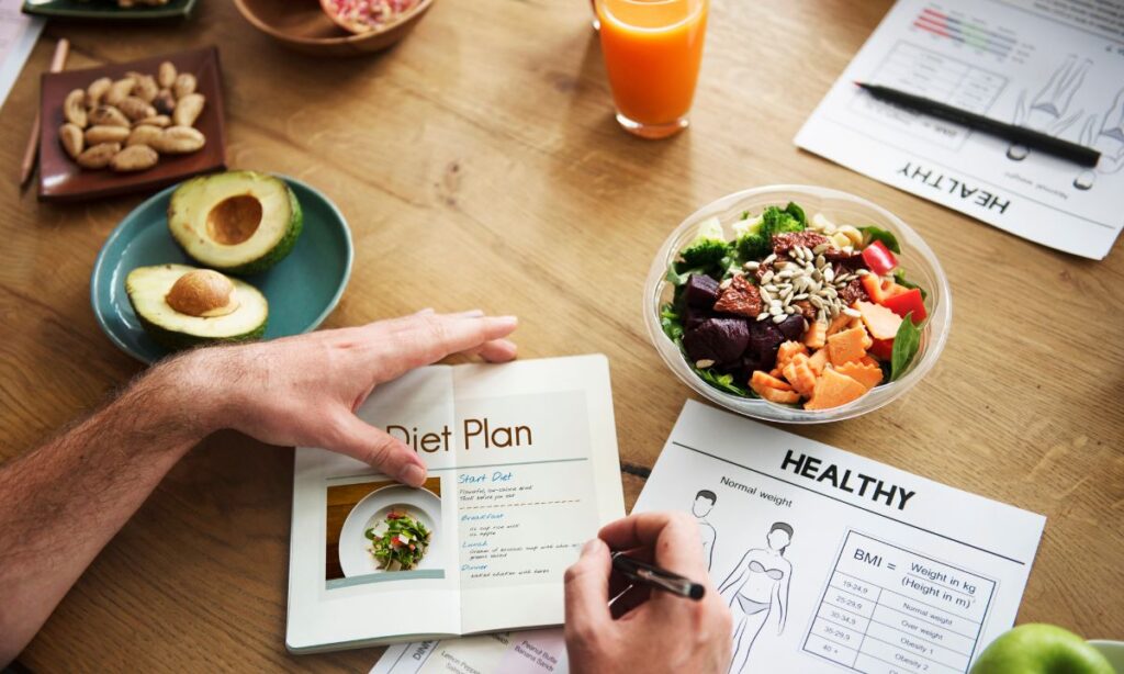 A table top view, there the hands holding a notebook with text written Diet plan. There are also a plate of food suitable for weight loss, e.g. avocado, nuts, pomegranate, salad and glass of orange juice. 