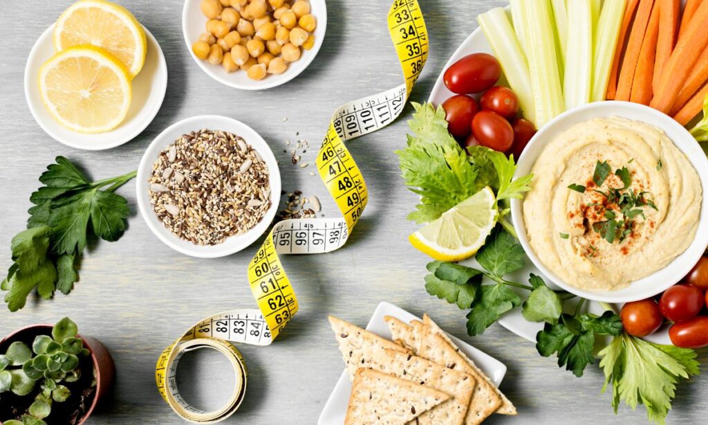 A top view of table full with plate of healthy diet suits for weight loss, e.g. slices of lemon, chickpeas, sesames, digestive biscuits, a hummus with vegetable sticks for dipping. Also a measuring tape lays on the middle.
